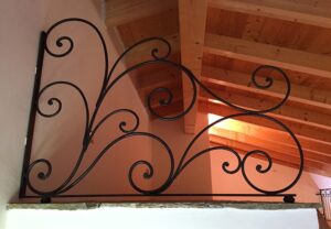 Ringhiera decorativa in ferro battuto nero con montante verticale, volute e riccioli sotto un soffitto in travi di legno