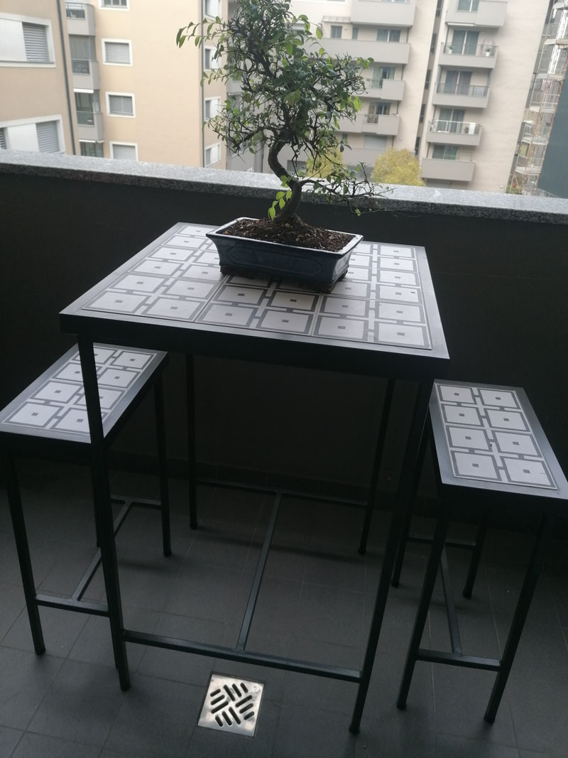 Tavolino alto e due sgabelli in ferro nero con piani in piastrelle geometriche grigie e bianche su un balcone